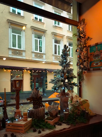 A cozy indoor scene features a decorative window display with various Christmas-themed items, including a small frosted tree adorned with blue lights, pinecones, greenery, and rustic wooden elements. Outside, through the window, a view of a street with a decorated building is visible.