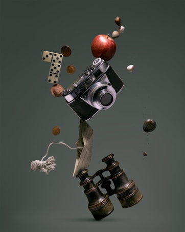 A surreal arrangement of various objects is floating in mid-air against a muted green background. The central focus is a vintage camera surrounded by a red apple, a pair of domino tiles, coins, a paper boat made from printed text, a ball of string, and a pair of old binoculars. Small pebbles and a couple of nuts appear to be suspended along with these items.