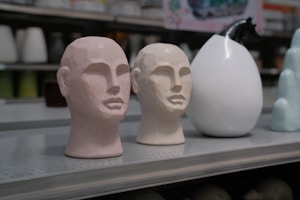 Two sculpted head busts with minimalist features are displayed on a metal shelf. The busts have a textured surface, one in a pinkish hue and the other in cream, positioned near a glossy, rounded white ceramic object and a light blue abstract sculpture. The setting appears to be a store or showroom display.