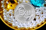 A silver coin labeled 'EXU' is surrounded by shiny pearls and two large gemstones, one yellow and one blue. The pearls are arranged around the coin, creating a luxurious and opulent display.