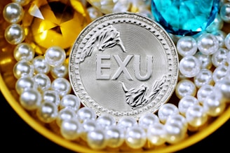 A silver coin labeled 'EXU' is surrounded by shiny pearls and two large gemstones, one yellow and one blue. The pearls are arranged around the coin, creating a luxurious and opulent display.