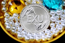 A silver coin labeled 'EXU' is surrounded by shiny pearls and two large gemstones, one yellow and one blue. The pearls are arranged around the coin, creating a luxurious and opulent display.