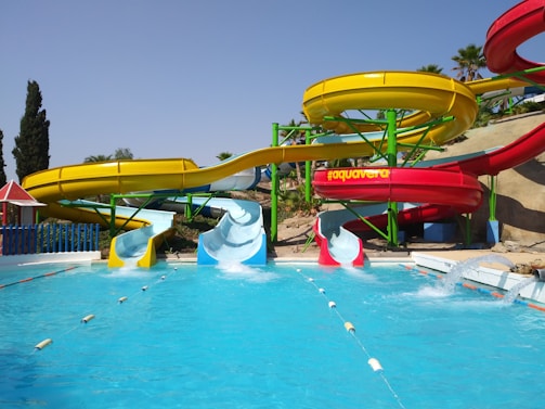 A vibrant water park features colorful water slides positioned above a clear blue pool. The slides are painted in bright yellow and red, with green support structures. Palm trees are visible in the background, adding a tropical feel to the setting. The pool has two diving sections with water splashing, suggesting activity and fun.