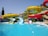 A vibrant water park features colorful water slides positioned above a clear blue pool. The slides are painted in bright yellow and red, with green support structures. Palm trees are visible in the background, adding a tropical feel to the setting. The pool has two diving sections with water splashing, suggesting activity and fun.