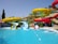 A vibrant water park features colorful water slides positioned above a clear blue pool. The slides are painted in bright yellow and red, with green support structures. Palm trees are visible in the background, adding a tropical feel to the setting. The pool has two diving sections with water splashing, suggesting activity and fun.