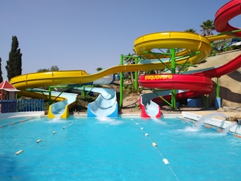 A vibrant water park features colorful water slides positioned above a clear blue pool. The slides are painted in bright yellow and red, with green support structures. Palm trees are visible in the background, adding a tropical feel to the setting. The pool has two diving sections with water splashing, suggesting activity and fun.