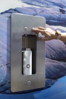 A hand wearing a watch is pressing a button on a stainless steel water dispenser mounted on a rocky textured background. Inside the dispenser, a white water bottle is being filled with liquid.