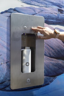 A hand wearing a watch is pressing a button on a stainless steel water dispenser mounted on a rocky textured background. Inside the dispenser, a white water bottle is being filled with liquid.
