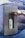 A hand wearing a watch is pressing a button on a stainless steel water dispenser mounted on a rocky textured background. Inside the dispenser, a white water bottle is being filled with liquid.