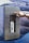 A hand wearing a watch is pressing a button on a stainless steel water dispenser mounted on a rocky textured background. Inside the dispenser, a white water bottle is being filled with liquid.