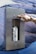 A hand wearing a watch is pressing a button on a stainless steel water dispenser mounted on a rocky textured background. Inside the dispenser, a white water bottle is being filled with liquid.