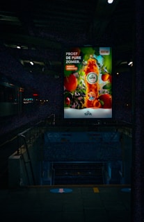 A brightly lit advertisement for a beverage brand featuring a bottle of mango apricot flavor is prominently displayed against a colorful fruit background. This advertisement is situated in a dimly lit urban environment, likely a train station, with visible stairways leading downwards and some faint signage.