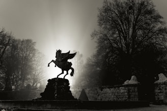 A silhouette of a Pegasus statue is prominently featured against a hazy, glowing background. The scene is framed by bare trees, which add to the dramatic and mystical atmosphere. The light source behind the statue creates a striking contrast with the dark foreground, highlighting the contours of the Pegasus.