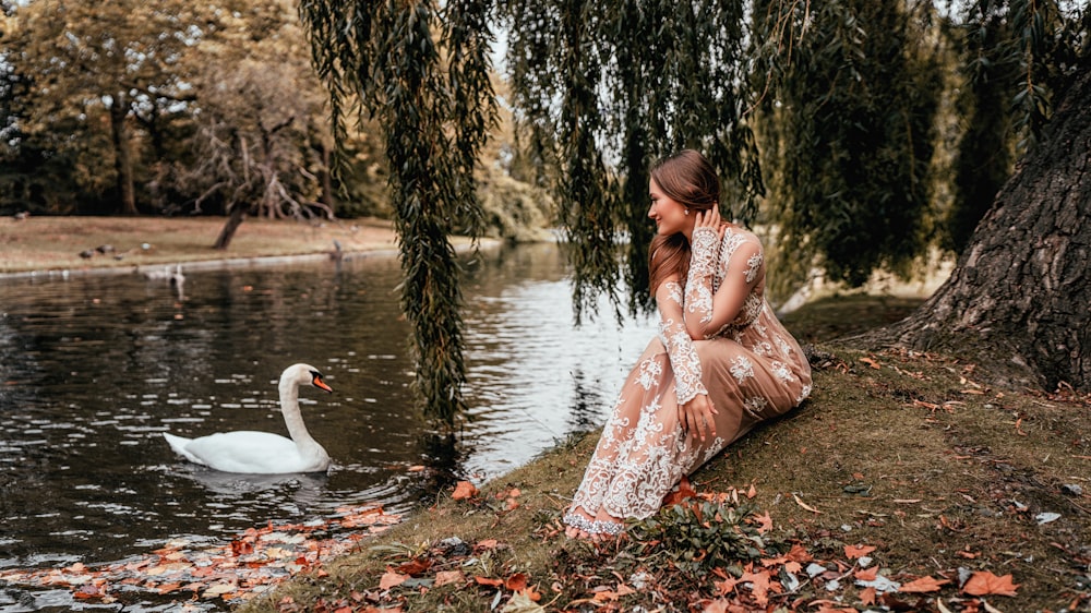 woman in pink dress sitting on ground with white swan on water during daytime