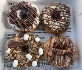 A quartet of donuts with rich toppings arranged neatly in a box on a newspaper-style lining. One donut is glazed with white icing and topped with chocolate drizzle and brownie bits. Another features chocolate icing with small marshmallows and a Hershey's chocolate piece. A third donut is adorned with chocolate and nut pieces, while the last one has a chocolate drizzle with pieces of waffle cone.