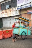 A street vendor wearing a red shirt and cap stands beside a brightly colored pushcart. The pushcart is filled with bottles and snacks and is painted in teal with yellow and orange accents. The street is bordered with orange traffic barriers, and in the background, there are weathered advertising signs on a building.