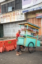 A street vendor wearing a red shirt and cap stands beside a brightly colored pushcart. The pushcart is filled with bottles and snacks and is painted in teal with yellow and orange accents. The street is bordered with orange traffic barriers, and in the background, there are weathered advertising signs on a building.