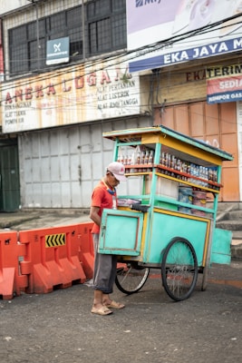 A street vendor wearing a red shirt and cap stands beside a brightly colored pushcart. The pushcart is filled with bottles and snacks and is painted in teal with yellow and orange accents. The street is bordered with orange traffic barriers, and in the background, there are weathered advertising signs on a building.