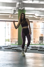 A woman wearing athletic clothing is dancing or exercising in an indoor gym setting. The background includes gym equipment and large windows that let in natural light. The atmosphere suggests movement and fitness.