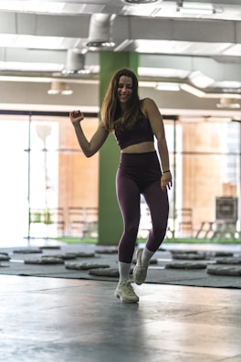 A woman wearing athletic clothing is dancing or exercising in an indoor gym setting. The background includes gym equipment and large windows that let in natural light. The atmosphere suggests movement and fitness.