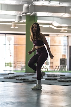 A woman is exercising indoors, wearing a dark sports outfit and sneakers. The spacious gym has a high ceiling with visible air ducts and large windows, allowing natural light to fill the room. Weight mats are spread across the floor.