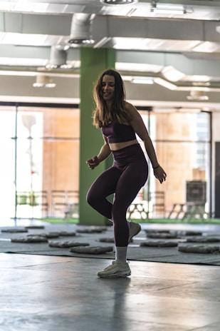 A woman is exercising indoors, wearing a dark sports outfit and sneakers. The spacious gym has a high ceiling with visible air ducts and large windows, allowing natural light to fill the room. Weight mats are spread across the floor.