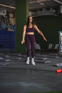 A woman in athletic wear is standing in a room with green walls. She appears to be smiling and in motion, suggesting she is either jumping or landing softly. The floor is covered with gray mats, and there is a mural on one wall depicting a comic book style character. In the foreground, an orange foam roller lies on the floor.