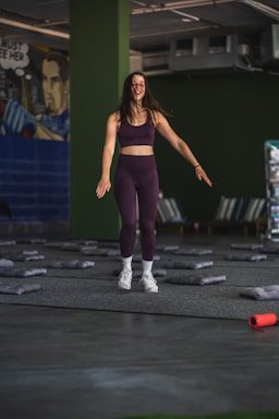 A woman in athletic wear is standing in a room with green walls. She appears to be smiling and in motion, suggesting she is either jumping or landing softly. The floor is covered with gray mats, and there is a mural on one wall depicting a comic book style character. In the foreground, an orange foam roller lies on the floor.