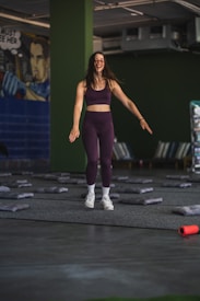 A woman in athletic wear is standing in a room with green walls. She appears to be smiling and in motion, suggesting she is either jumping or landing softly. The floor is covered with gray mats, and there is a mural on one wall depicting a comic book style character. In the foreground, an orange foam roller lies on the floor.