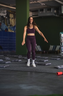 A woman in athletic wear is standing in a room with green walls. She appears to be smiling and in motion, suggesting she is either jumping or landing softly. The floor is covered with gray mats, and there is a mural on one wall depicting a comic book style character. In the foreground, an orange foam roller lies on the floor.