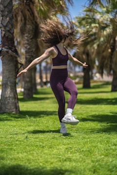 A person in athletic wear performs a joyful dance or exercise on a grassy field surrounded by palm trees. The scene is bright and sunny, and the person's hair flows dynamically with their movement.