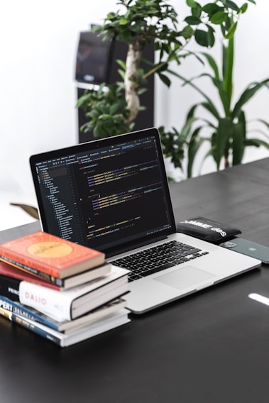 A workspace featuring a laptop displaying code, accompanied by a stack of books. The background includes green plants, creating a fresh and vibrant atmosphere. The desk also has a smartphone and a closed notebook.