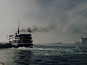 A large ferry with a flag on top emits dark smoke as it moves through a body of water. In the background, another smaller ship is seen on the horizon, with a building on the right side of the image. The sky appears overcast, with cloud cover creating a muted atmosphere.