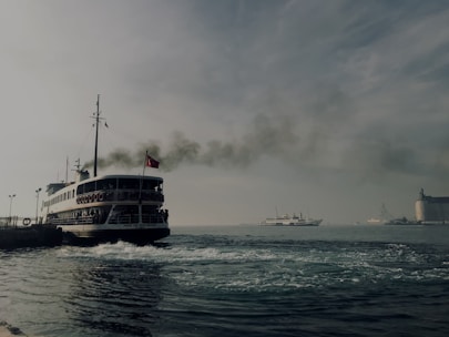 A large ferry with a flag on top emits dark smoke as it moves through a body of water. In the background, another smaller ship is seen on the horizon, with a building on the right side of the image. The sky appears overcast, with cloud cover creating a muted atmosphere.