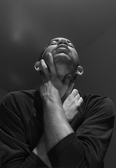 A person with an expression of anguish or distress has both hands clasped around their neck. The grayscale tones add to the dramatic effect, highlighting the texture of the skin and the emotion conveyed through facial expression and posture.