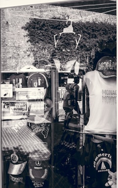 A shop display window featuring various Monaco-themed merchandise. Items include caps, t-shirts, and bags with prints related to Monaco. The window reflects the stone wall and plants outside, along with the silhouette of a person walking. Signs in French are visible, indicating a location in a French-speaking region.