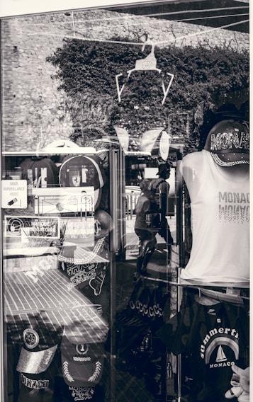 A shop display window featuring various Monaco-themed merchandise. Items include caps, t-shirts, and bags with prints related to Monaco. The window reflects the stone wall and plants outside, along with the silhouette of a person walking. Signs in French are visible, indicating a location in a French-speaking region.