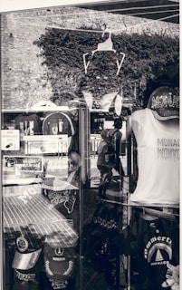 A shop display window featuring various Monaco-themed merchandise. Items include caps, t-shirts, and bags with prints related to Monaco. The window reflects the stone wall and plants outside, along with the silhouette of a person walking. Signs in French are visible, indicating a location in a French-speaking region.