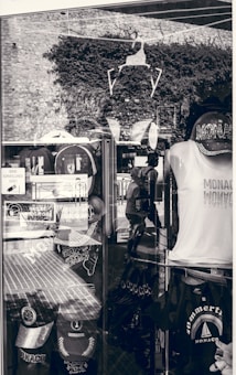 A shop display window featuring various Monaco-themed merchandise. Items include caps, t-shirts, and bags with prints related to Monaco. The window reflects the stone wall and plants outside, along with the silhouette of a person walking. Signs in French are visible, indicating a location in a French-speaking region.