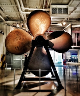 A large metallic propeller is displayed prominently indoors, mounted on a sturdy stand. The environment appears to be an industrial or museum setting with various technical exhibits in the background. Lighting is a mix of natural light from a large window and artificial ceiling lights. A person is visible near the propeller, providing a sense of scale.