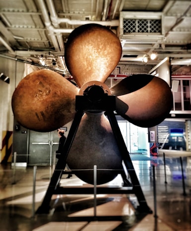 A large metallic propeller is displayed prominently indoors, mounted on a sturdy stand. The environment appears to be an industrial or museum setting with various technical exhibits in the background. Lighting is a mix of natural light from a large window and artificial ceiling lights. A person is visible near the propeller, providing a sense of scale.