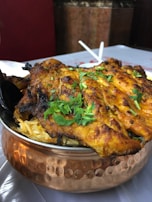 A serving of grilled, spiced chicken placed atop a bed of rice in a traditional copper bowl, garnished with fresh cilantro. The dish is likely an Indian or Middle Eastern delicacy, with vibrant colors and an appetizing presentation.