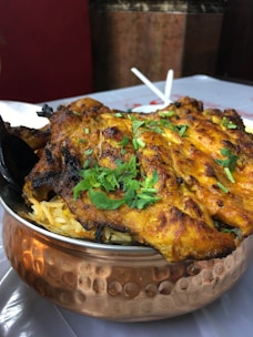 A serving of grilled, spiced chicken placed atop a bed of rice in a traditional copper bowl, garnished with fresh cilantro. The dish is likely an Indian or Middle Eastern delicacy, with vibrant colors and an appetizing presentation.