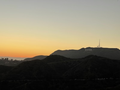 A scenic landscape depicts the Hollywood Hills at sunset with the iconic Hollywood sign visible on the hillside. The sky is a gradient of warm colors transitioning from orange near the horizon to a light blue as it extends upward. The silhouette of the hills is prominently featured in the foreground.