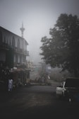 A misty street scene featuring a car with an open hood on the road, surrounded by buildings with traditional architecture and a tall minaret. Several people are gathered near a shop, partially obscured by fog. A large tree adds to the serene, quiet atmosphere.