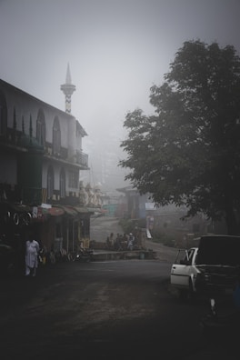 A misty street scene featuring a car with an open hood on the road, surrounded by buildings with traditional architecture and a tall minaret. Several people are gathered near a shop, partially obscured by fog. A large tree adds to the serene, quiet atmosphere.