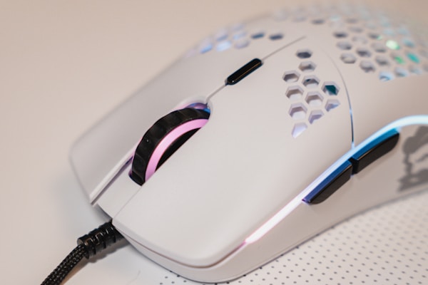 A close-up view of a white computer mouse with a perforated honeycomb design and RGB lighting. The mouse features side buttons and a scroll wheel, connected by a braided cable. Its sleek and modern aesthetic is complemented by subtle color-changing light effects.