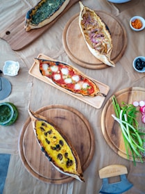 A selection of four pide breads is arranged on wooden serving boards. Each pide is topped with different ingredients: one with spinach, another with cheese and red onions, a third with a tomato and mozzarella combination, and the fourth featuring a creamy, possibly egg-based topping with mushrooms. Surrounding the pides are small bowls with olives, dips, and a side of fresh vegetables like scallions and radishes.