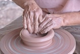 Hands of a potter working clay on a spinning pottery wheel, shaping a clay bowl. The hands are covered in wet clay, indicating active engagement in the pottery-making process, with a focus on craftsmanship and artistry.
