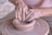 Hands of a potter working clay on a spinning pottery wheel, shaping a clay bowl. The hands are covered in wet clay, indicating active engagement in the pottery-making process, with a focus on craftsmanship and artistry.